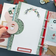 a hand holding a button on top of an open christmas card book with other cards and decorations around it