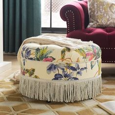 a floral ottoman with tassels on it in front of a red chair and window