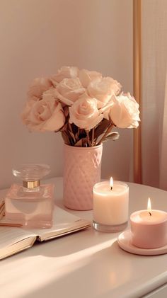 white roses in a pink vase and two candles on a small table with a book