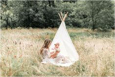 a woman holding a baby sitting in a field next to a teepee with trees behind her