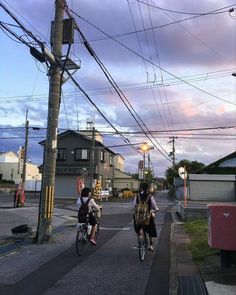 two people riding bikes down the street with power lines above them and houses in the background