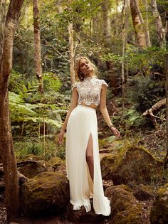 a woman standing in the woods wearing a white skirt and crop top with high slit