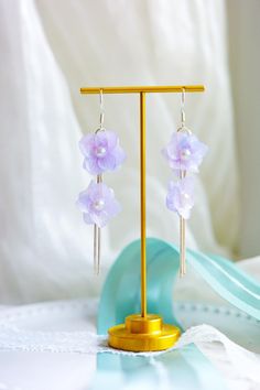 purple flowers are hanging from earrings on a gold plated stand with white fabric in the background