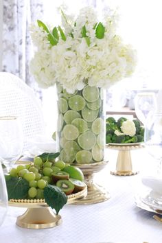 white flowers and green grapes in a vase on a table with other plates, silverware and glassware