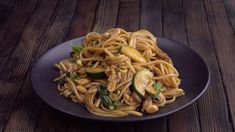 a black plate topped with pasta and vegetables on top of a wooden table next to a fork