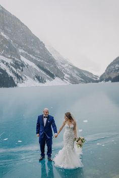 a bride and groom holding hands in front of a lake surrounded by snow covered mountains