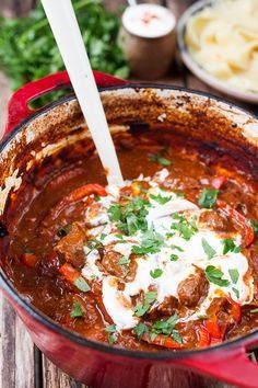 a red pot filled with meat and sauce on top of a wooden table next to pasta