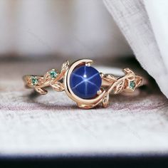 a gold ring with a blue stone and green stones on the side, sitting on top of a piece of cloth