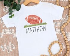 Elevate your little one's style game with our Toddler and Kid Football Shirt, a winning combination of sporty flair and personalized charm. Designed for comfort and individuality, this shirt features a prominent football graphic and can be customized with your child's name, creating a unique piece that's perfect for playtime, outings, and more. 🏈 Sporty Elegance: Our shirt showcases a meticulously designed football, capturing the excitement of the game in a stylish and kid-friendly way. The graphic adds a touch of athleticism to your child's wardrobe, sure to catch the eye of fellow sports enthusiasts. 👦👧 Perfect Fit for Toddlers and Kids: Available in a range of sizes to accommodate toddlers and older children alike, this shirt boasts a comfortable fit that allows for free movement dur Personalized White Sporty T-shirt, Personalized Cotton T-shirt For Game Day, Sporty Personalized T-shirt For Sports, Personalized Sporty T-shirt For Sports, Personalized Short Sleeve Tops For Sports Events, Sporty Personalized T-shirt For Sports Events, Sporty Personalized Tops For Sports Events, Personalized Sporty Tops For Sports, White Personalized Team Spirit Top