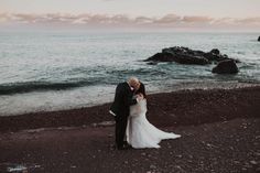 a bride and groom standing on the beach at sunset with their arms around each other