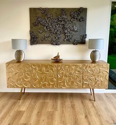 a golden sideboard with two lamps on it in front of a painting and wood flooring