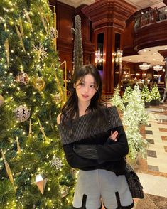 a woman standing in front of a christmas tree with her arms crossed and looking at the camera