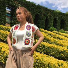 a woman standing in front of some bushes and yellow flowers with her hands on her hips