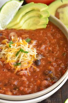 a white bowl filled with chili and cheese next to an avocado on the side