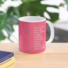 a pink coffee mug sitting on top of a wooden table next to a book and plant