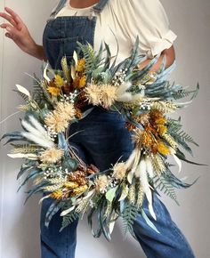 a woman wearing overalls and holding a wreath made out of dried flowers with leaves