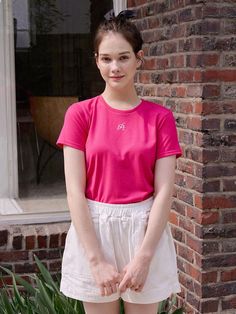 a woman standing in front of a brick building wearing white shorts and a pink shirt