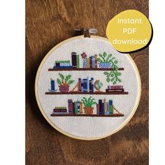 a cross - stitch book shelf with books and plants in it on a wooden surface