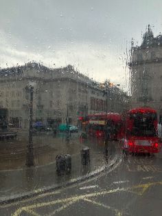 two red double decker buses driving down a rain soaked street next to tall buildings on a rainy day