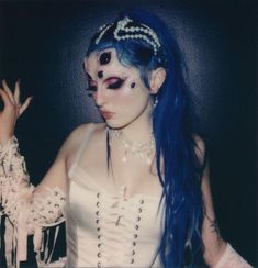 a woman with blue hair and makeup holding her hand up to the camera while wearing white clothing