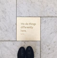 there is a sign that says we do things differently here on the marble floor next to a pair of black shoes