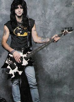 a man with long black hair holding a guitar in his right hand and posing for the camera