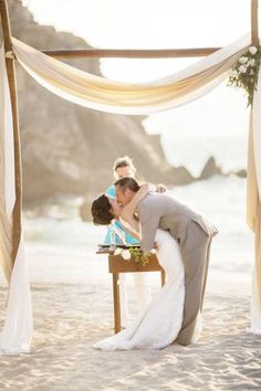 a bride and groom kissing on the beach under an arch decorated with white draping