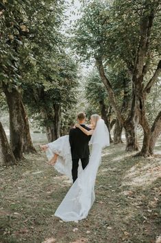 a bride and groom are walking through the woods in their wedding attire, holding each other's arms