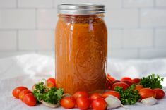 a glass jar filled with tomato sauce surrounded by tomatoes and parsley on a white cloth