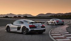 two silver sports cars driving on a race track at sunset with mountains in the background