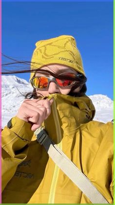 a woman in yellow jacket and red sunglasses standing on top of snow covered ground with mountains behind her