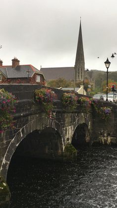 an old stone bridge with flowers growing on it and a church steeple in the background