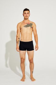 a shirtless man with tattoos standing in front of a white wall wearing black trunks