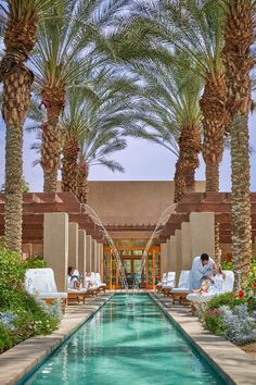 two people are sitting at the edge of a pool surrounded by palm trees