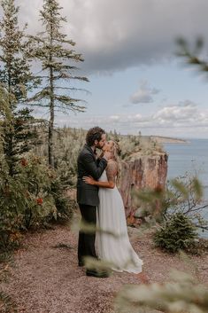 a bride and groom kissing in front of the ocean on their wedding day, surrounded by pine trees