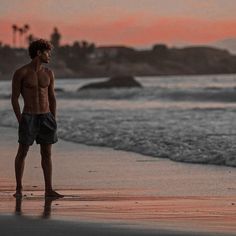 a man standing on top of a beach next to the ocean at sunset with his shirt off