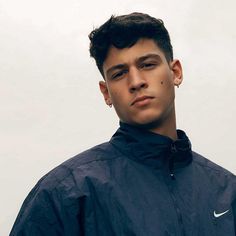 a young man wearing a nike jacket and ear piercings standing in front of a white wall