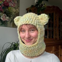 a woman wearing a knitted bear hat and neckwarmer in front of a potted plant