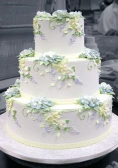 a three tiered white cake with blue flowers on it's side and people in the background