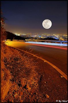 a full moon is seen above the city lights in this nighttime photo taken from an empty road