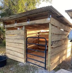 an outhouse made from pallets and wood with metal bars on the roof, next to a trash can