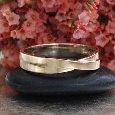 two gold wedding bands sitting on top of a black rock next to pink flower bush