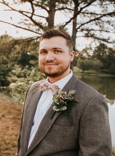 a man in a suit and bow tie standing next to a lake with trees behind him