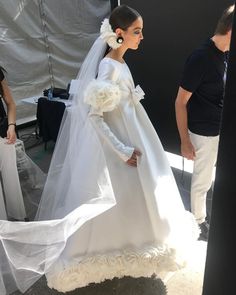 a woman in a white wedding gown and veil walking down the street with another man behind her