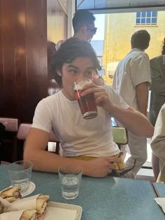 a young man sitting at a table drinking from a tall glass in front of him