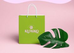 a green shopping bag next to a monster leaf on a pink background with the words kumiko written in white