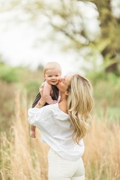 a woman holding a baby in her arms and kissing it's face while standing in tall grass