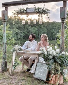 a man and woman sitting at a table in the middle of an outdoor area with flowers