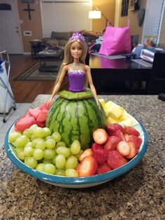 a doll sitting on top of a watermelon bowl filled with grapes and strawberries