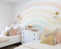 two white beds with pillows on them next to a wall painted with rainbow swirls